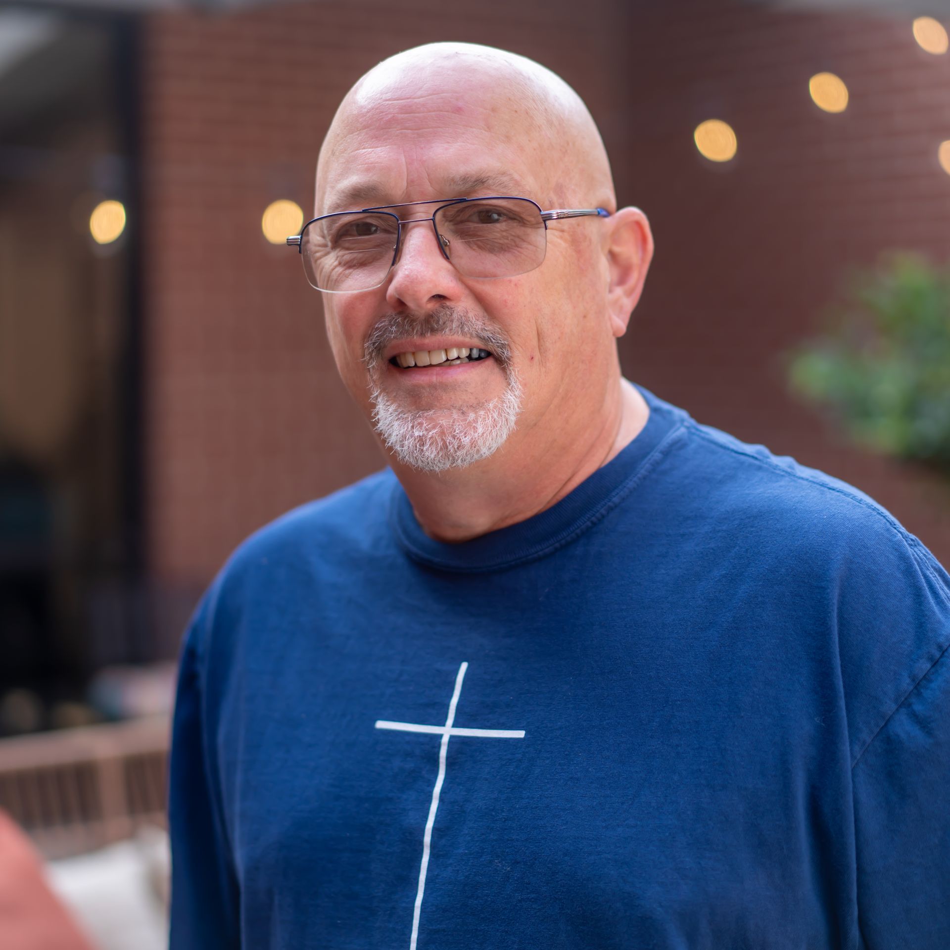 Bald man with glasses wearing a blue shirt with a white cross. Smiling outdoors near brick wall.