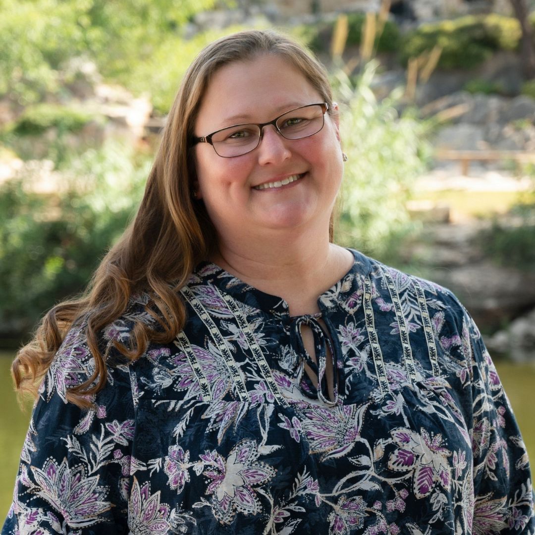 Bald man with glasses wearing a blue shirt with a white cross. Smiling outdoors near brick wall.