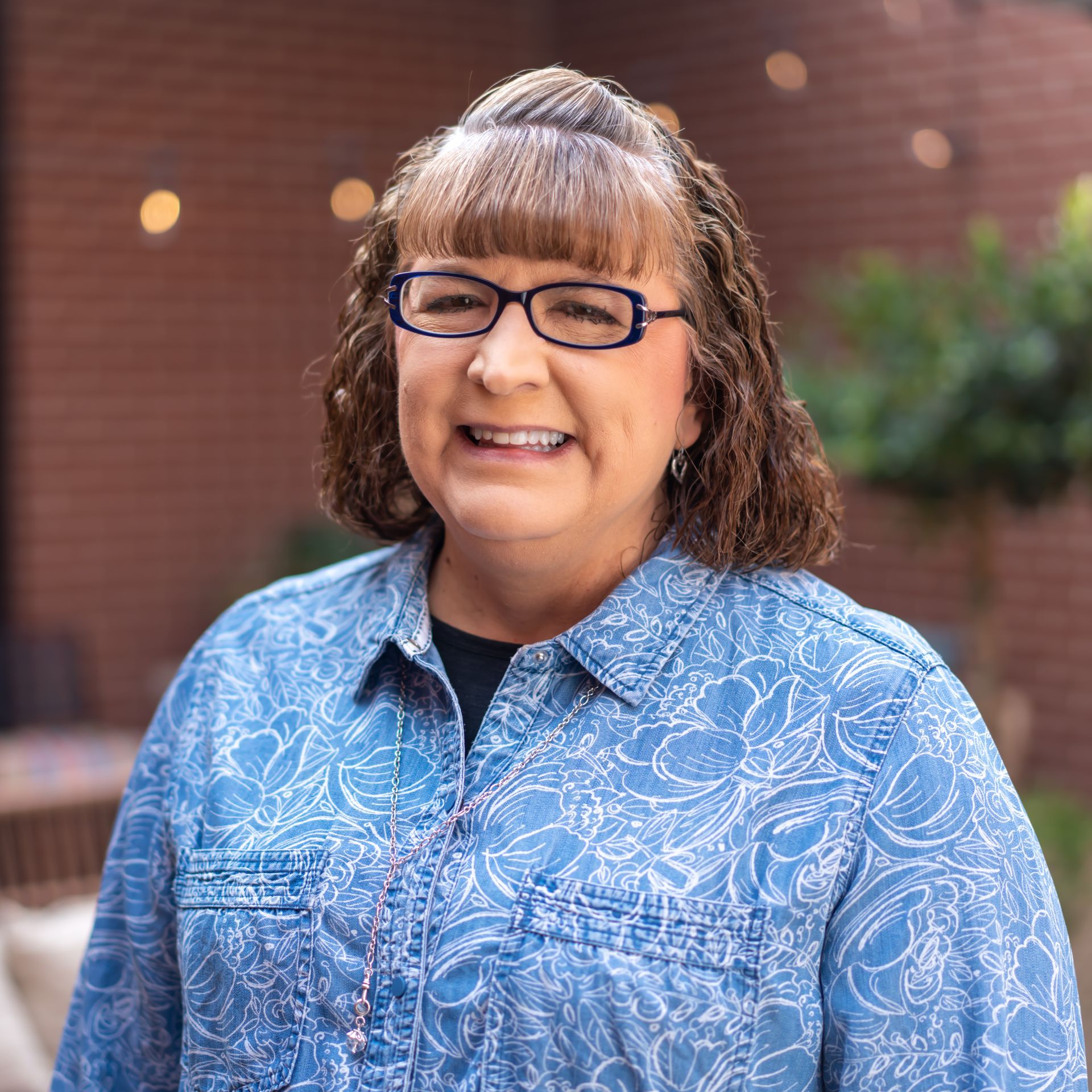 Woman with glasses smiling, wearing a blue floral shirt, standing outdoors in front of a brick wall.