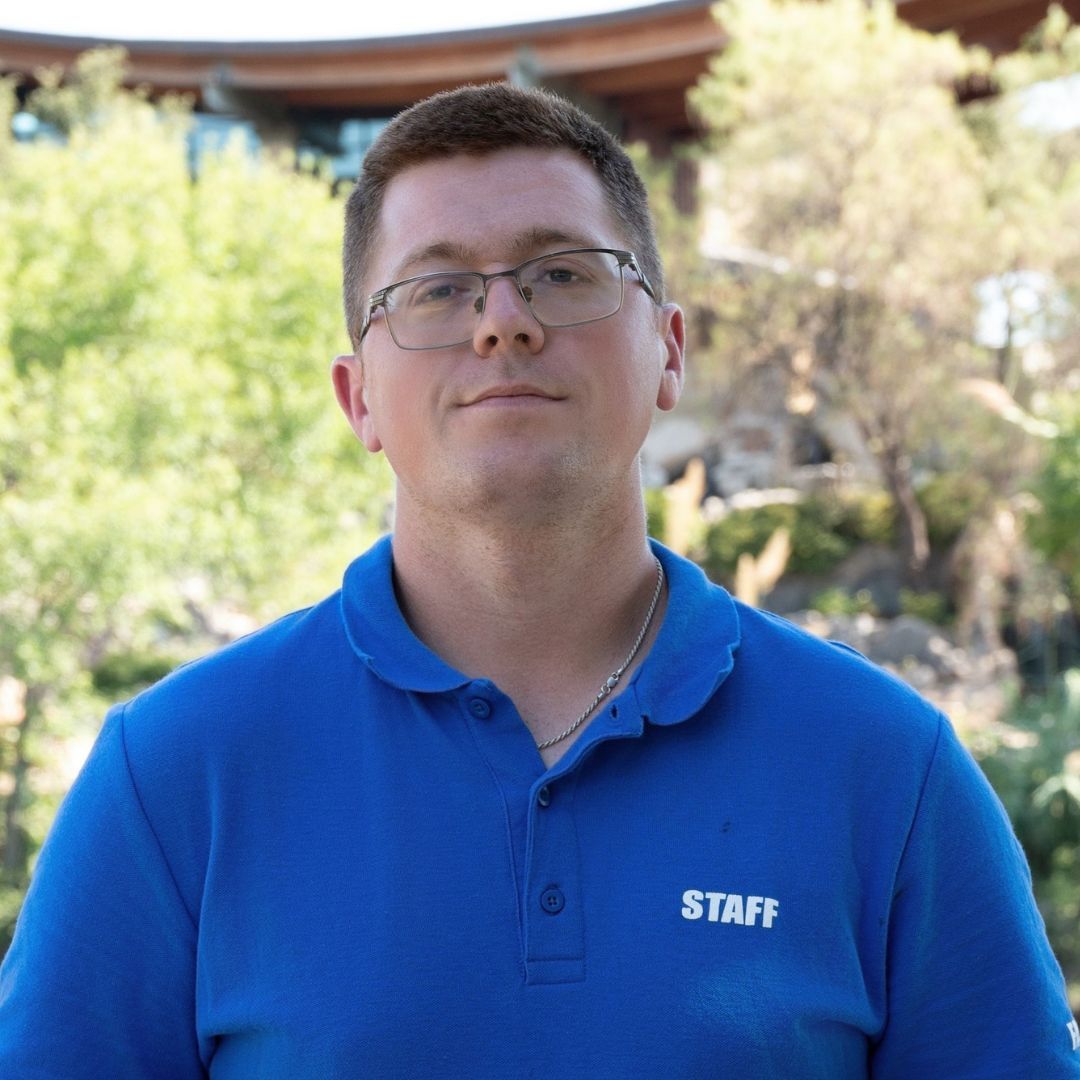 Man with glasses in blue floral shirt, looking off-camera. Outdoors, brick background.