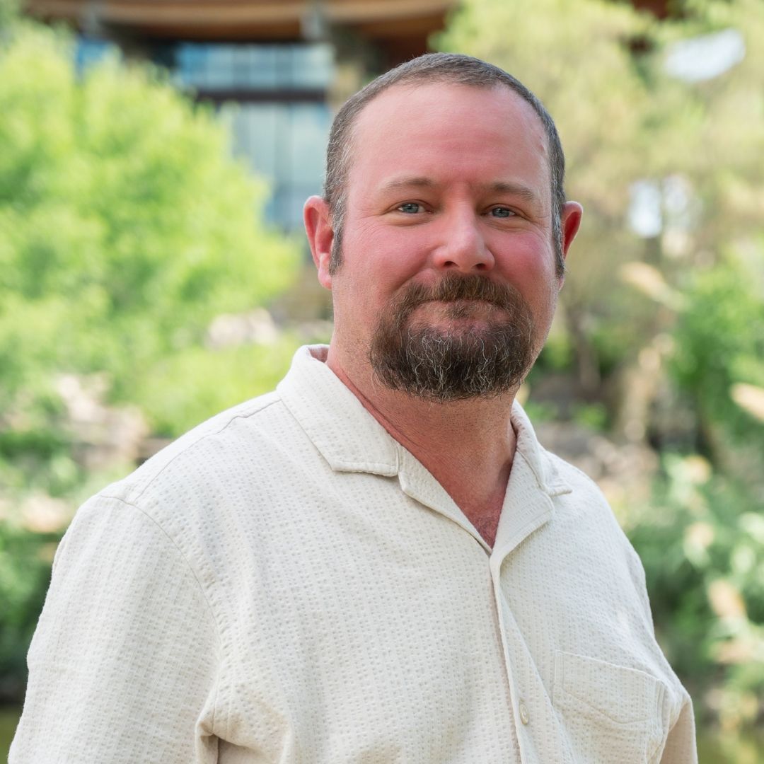 Man in blue blazer and white shirt smiles outdoors.