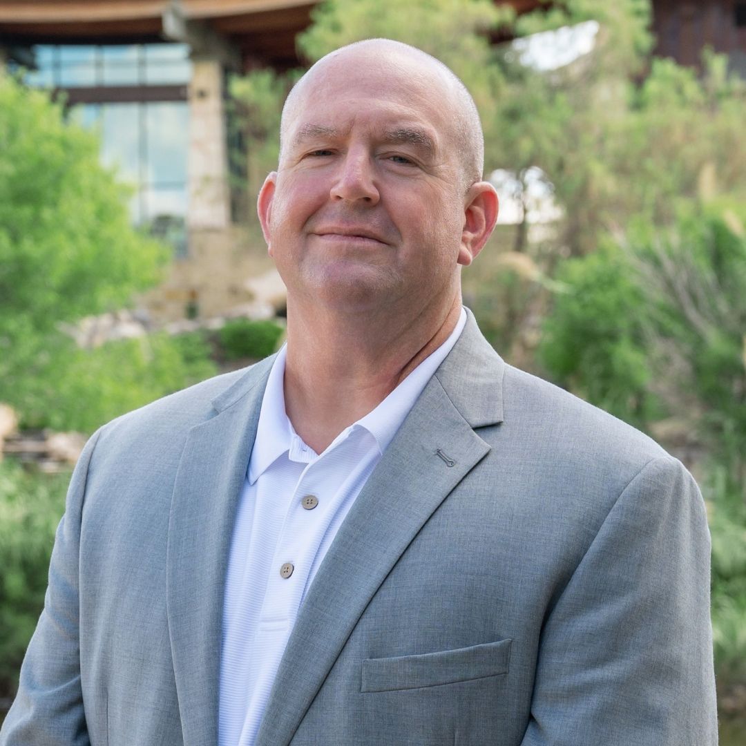 Man wearing a navy blazer and white polo smiles outdoors near a brick wall and string lights.