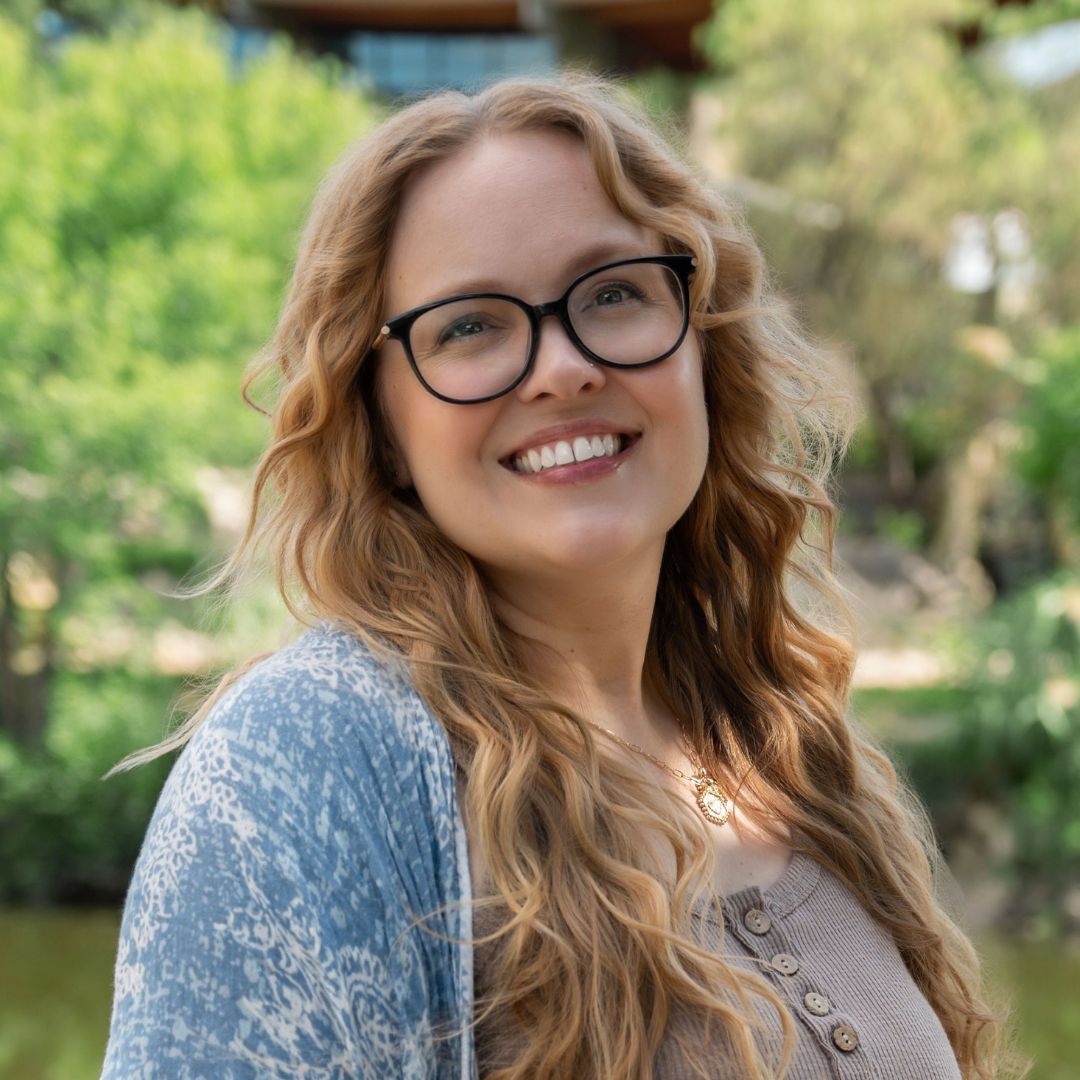 Woman with blonde hair and glasses smiles in front of a brick building.