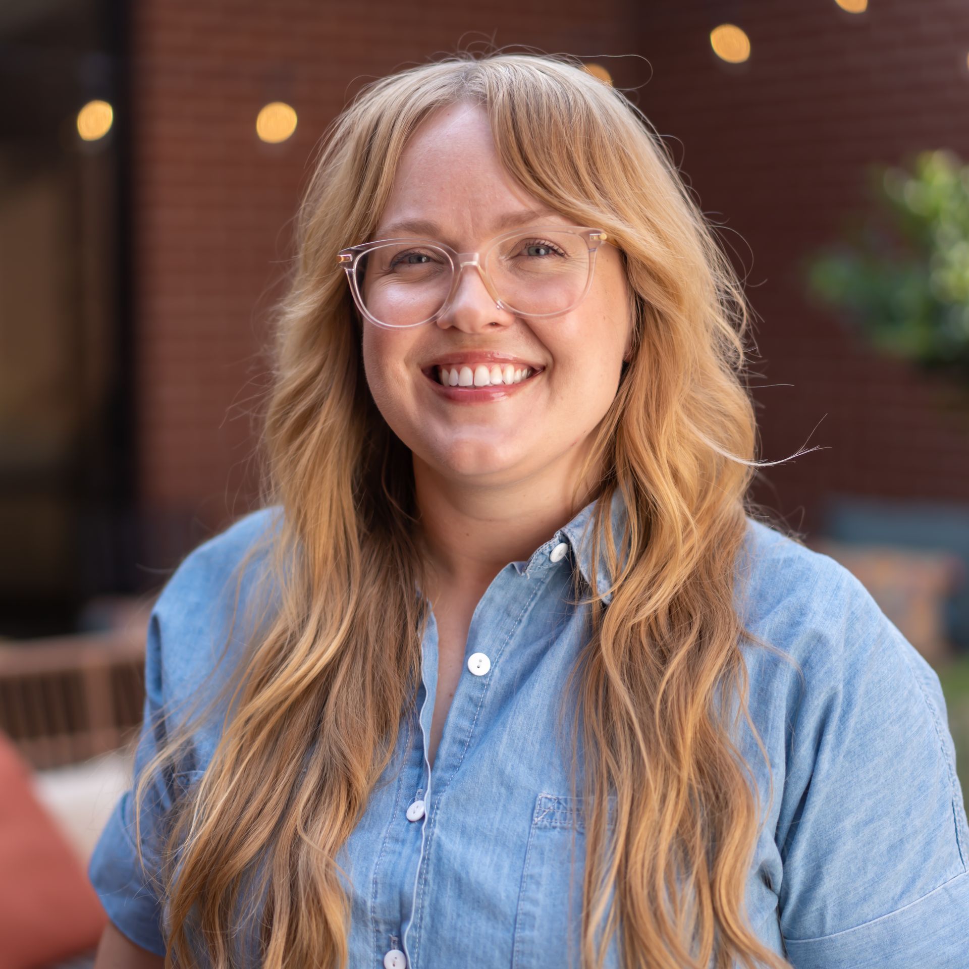 Woman with blonde hair and glasses smiles in front of a brick building.