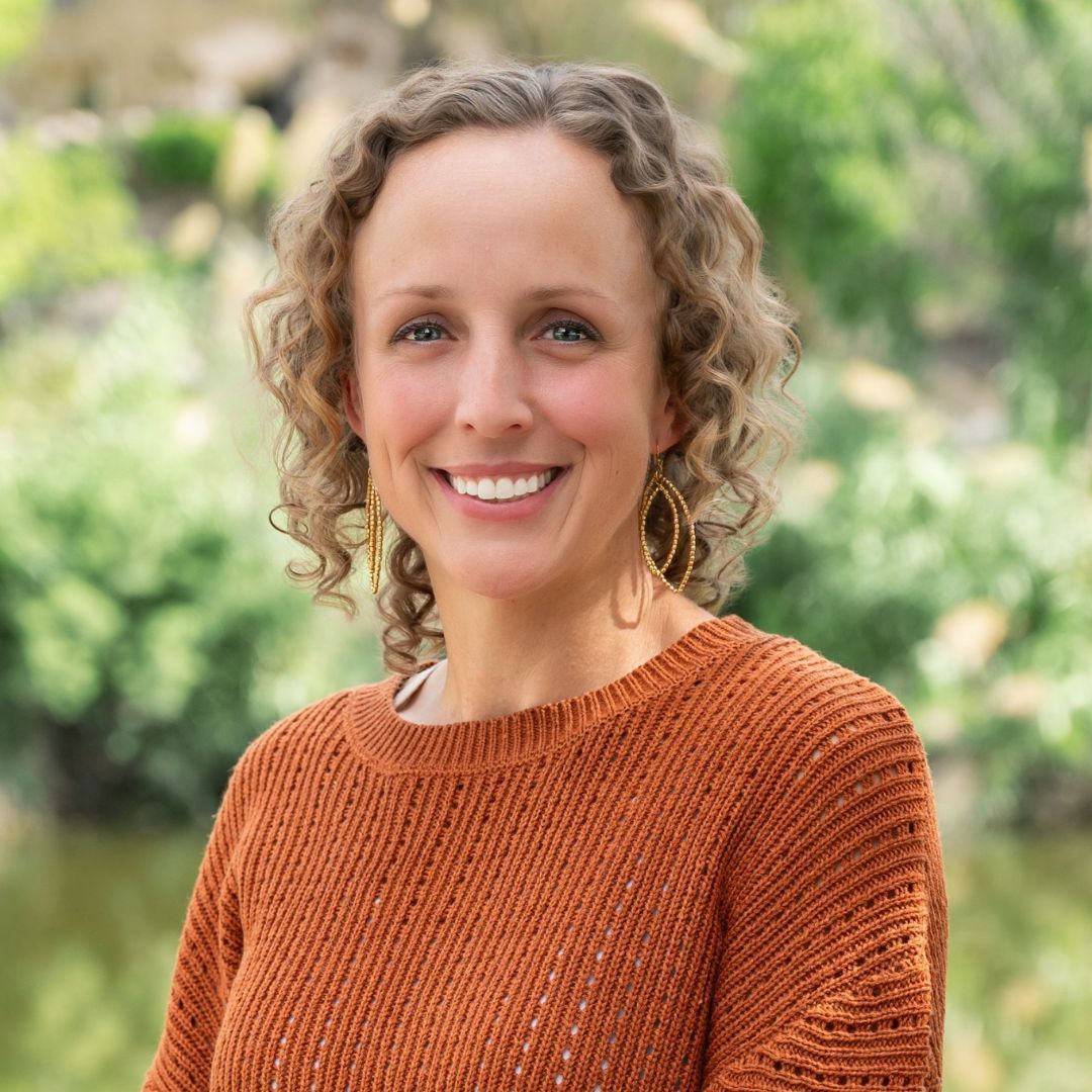 Bald man with glasses and beard, in a blue button-down shirt, smiles outdoors.