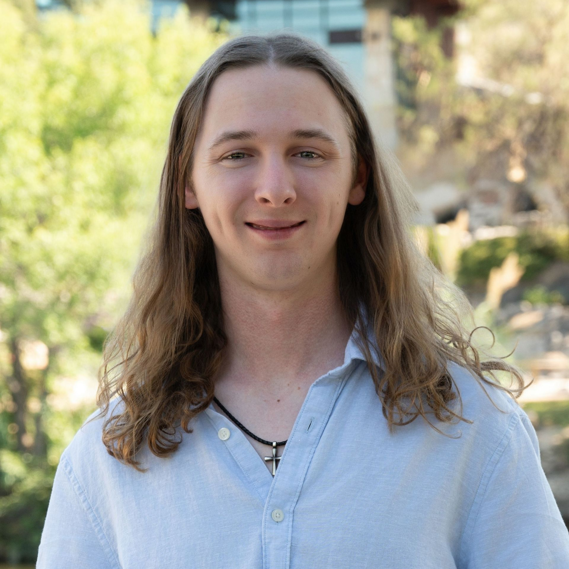 Man with light brown hair, smiling, wearing a gray polo shirt, standing outside with brick wall in the background.