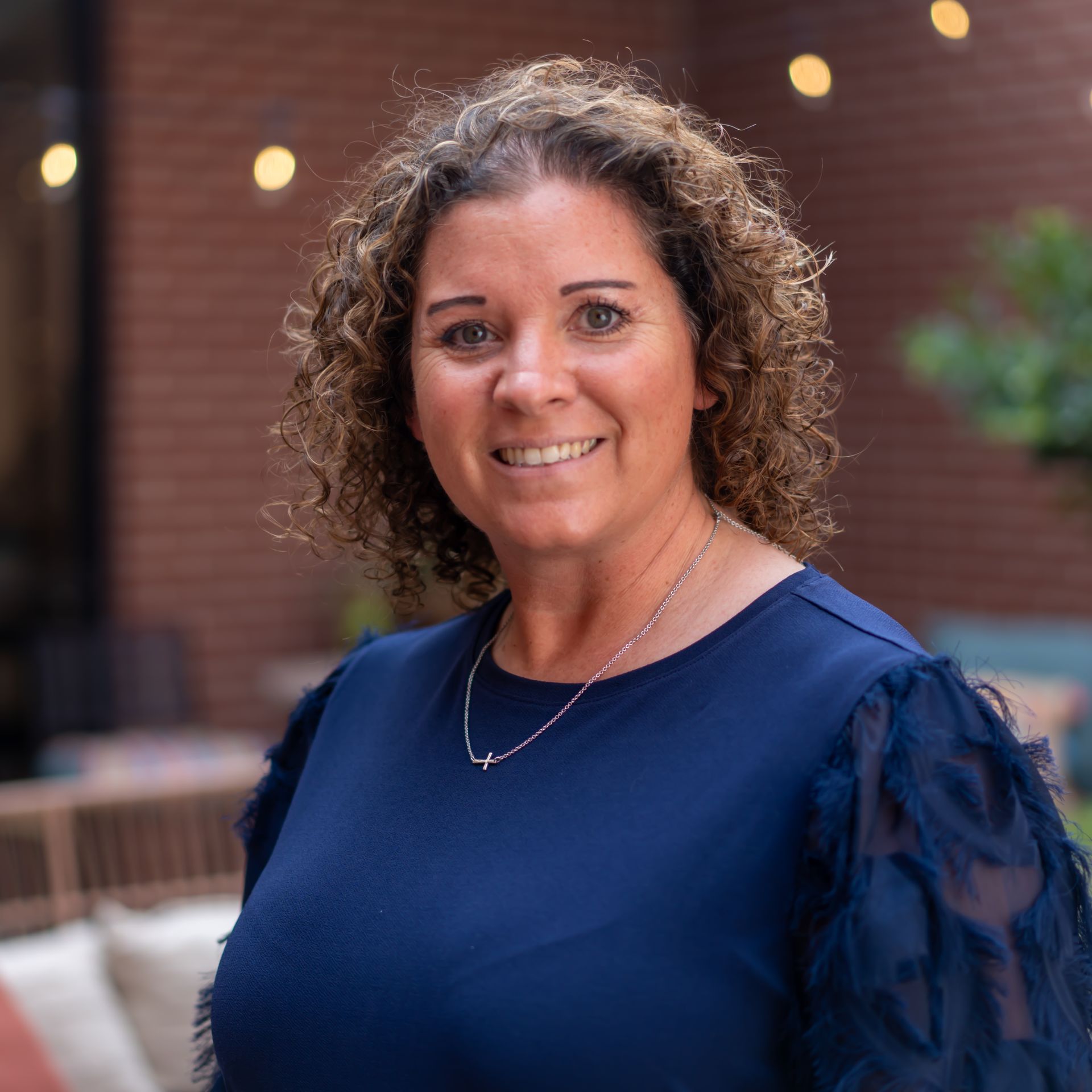 Woman in blue top smiles outdoors, brick backdrop, decorative sleeves.