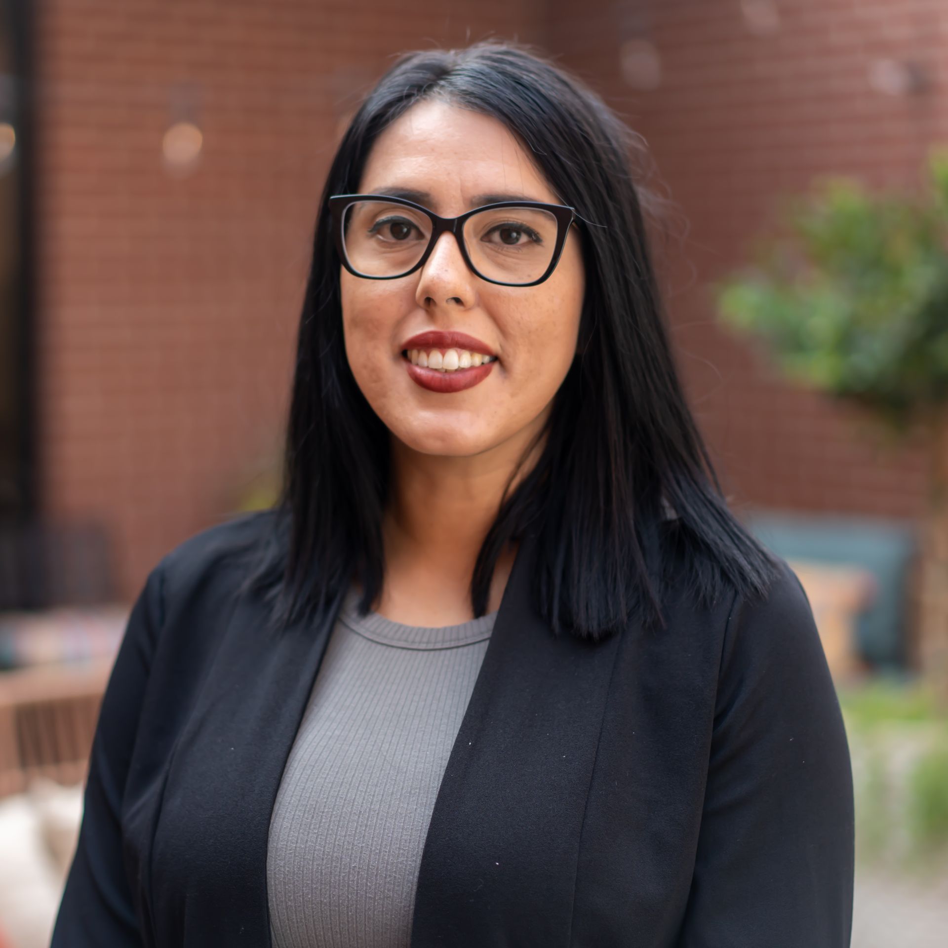 Woman wearing glasses and a black blazer smiles outdoors near a brick wall.