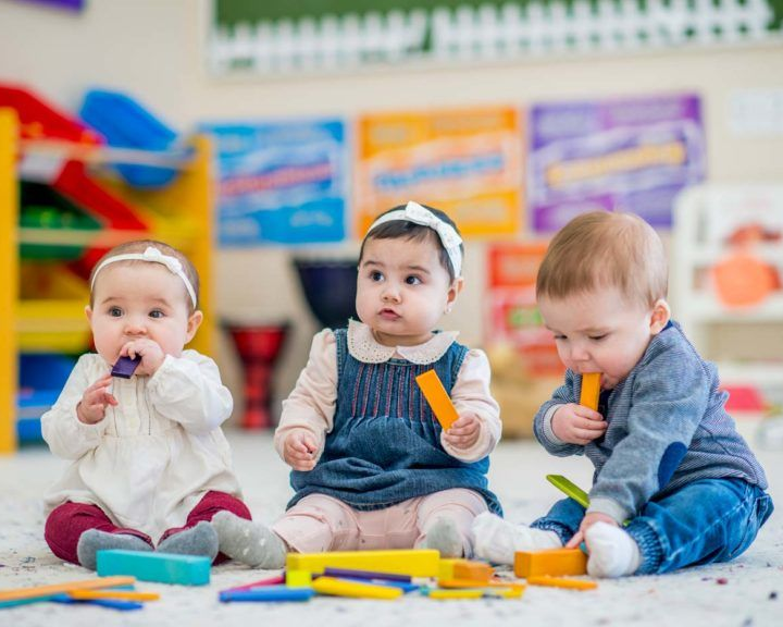 Three babies sit on a playroom floor, holding and chewing toys.