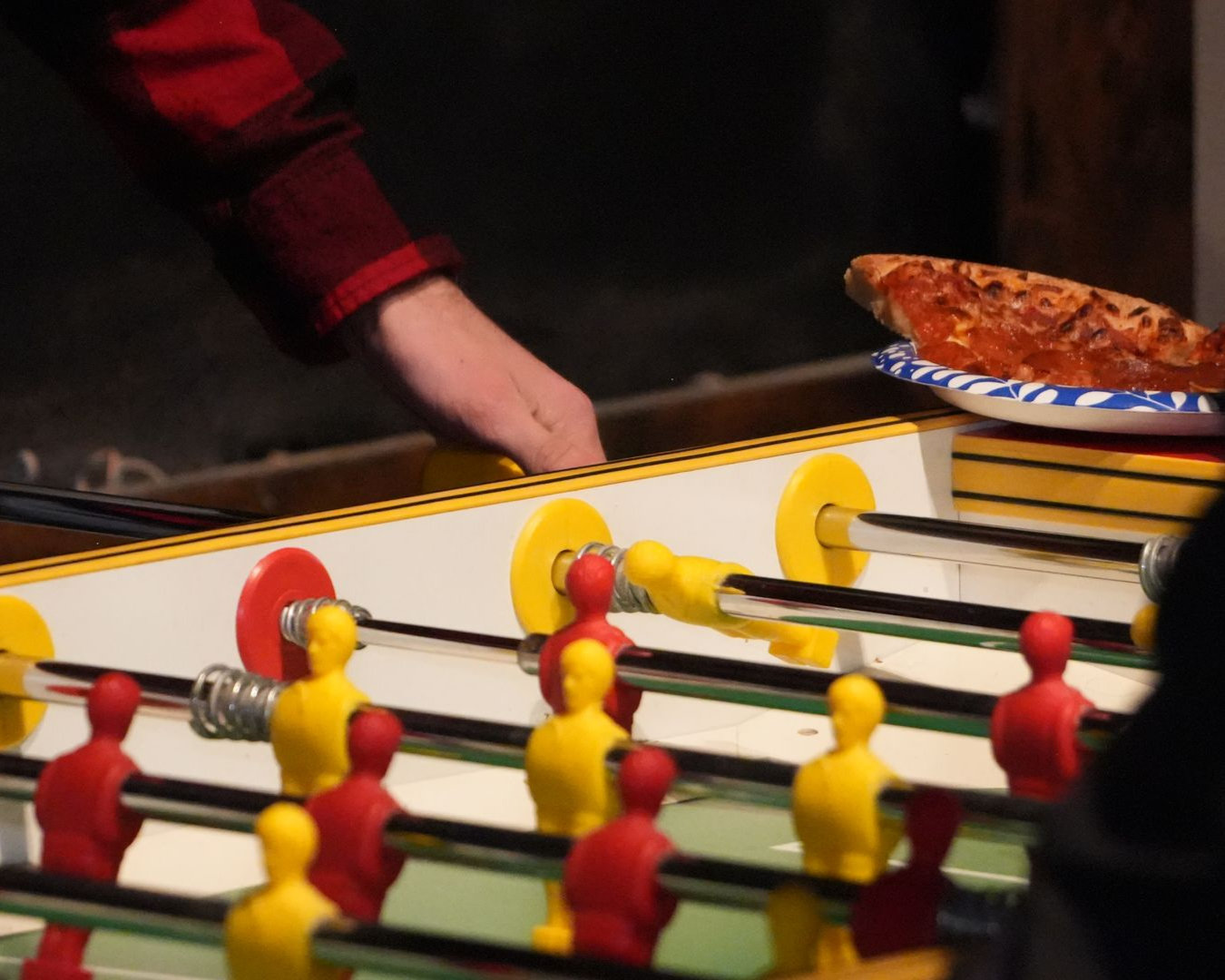 Foosball table with red and yellow players. A hand reaches toward the table, pizza slice nearby.