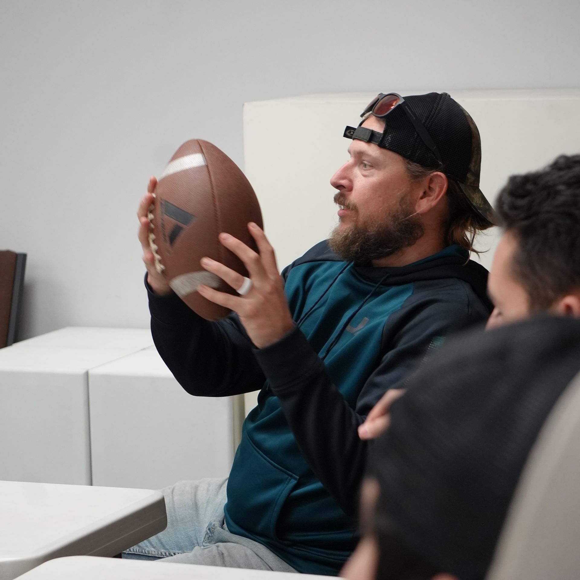 Man holding football, wearing a cap and blue hoodie, discussing with other people at a table.