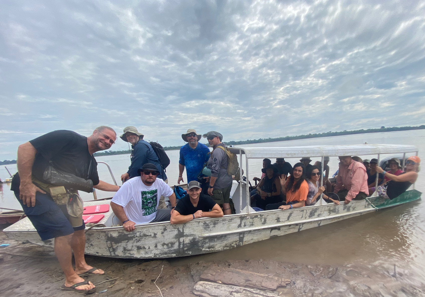 Group of people on a boat on a river, cloudy sky overhead. One man smiles, others look at the camera.
