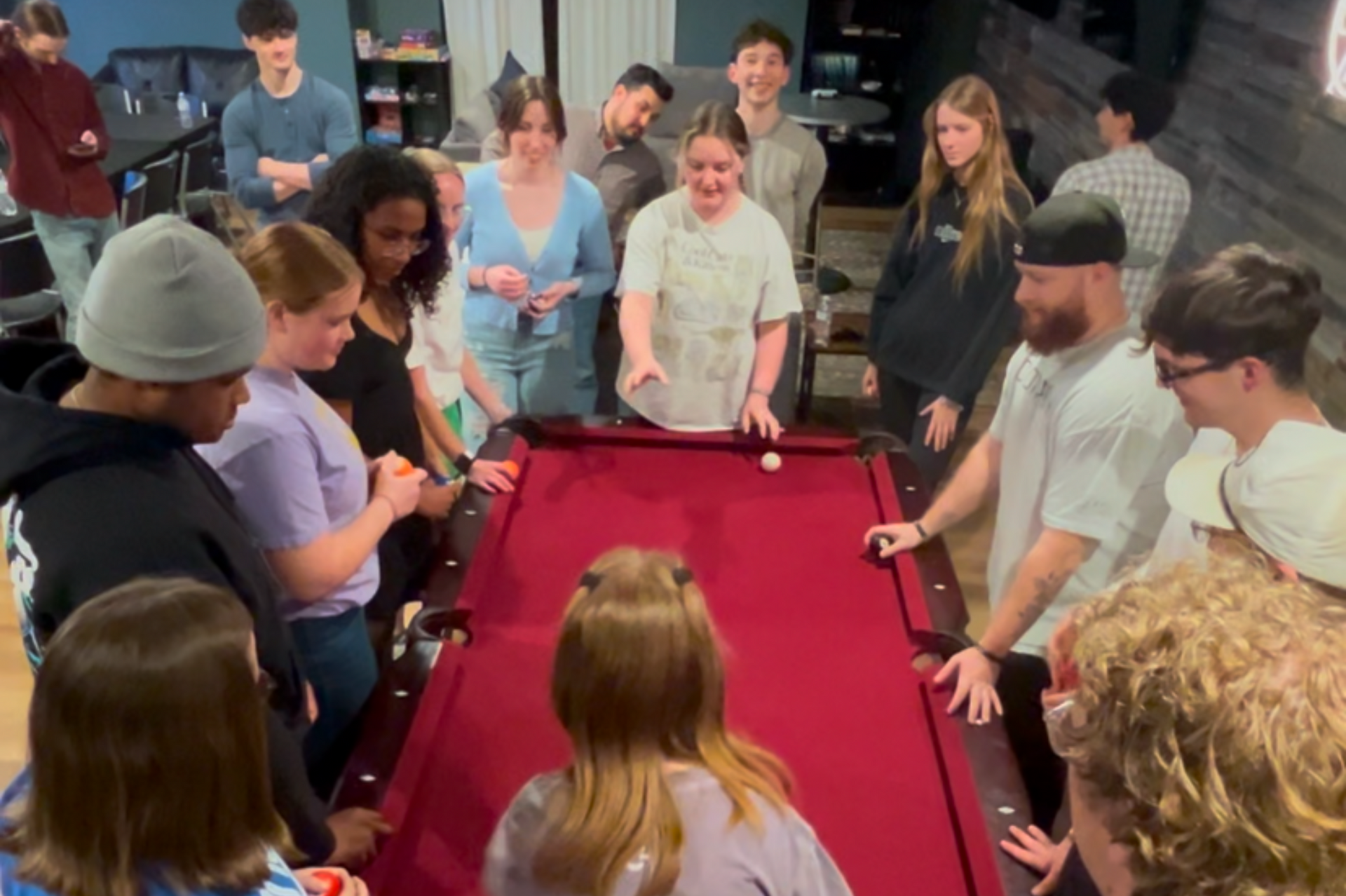 Group of people playing pool in a room with a red table, watching the white cue ball.