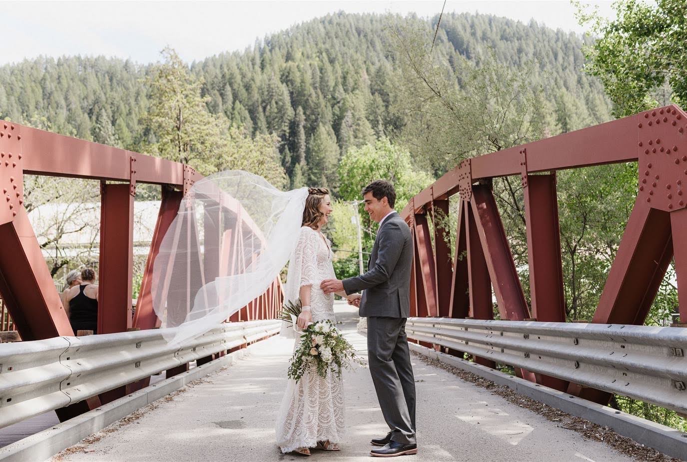 A bride and groom are standing on a bridge holding hands.