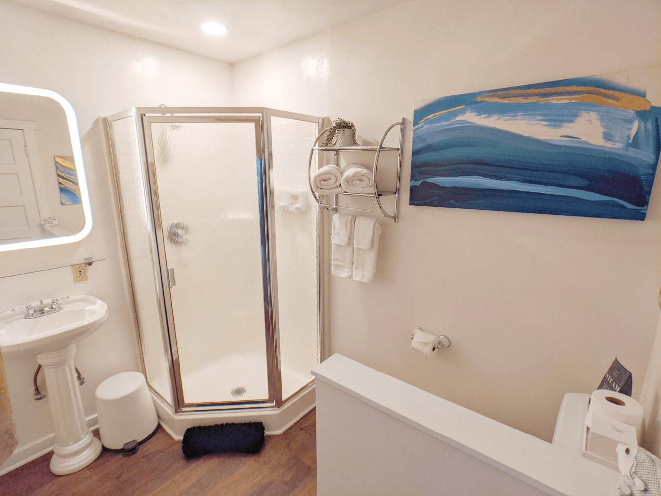 A bathroom with a walk in shower , sink , mirror and painting on the wall.