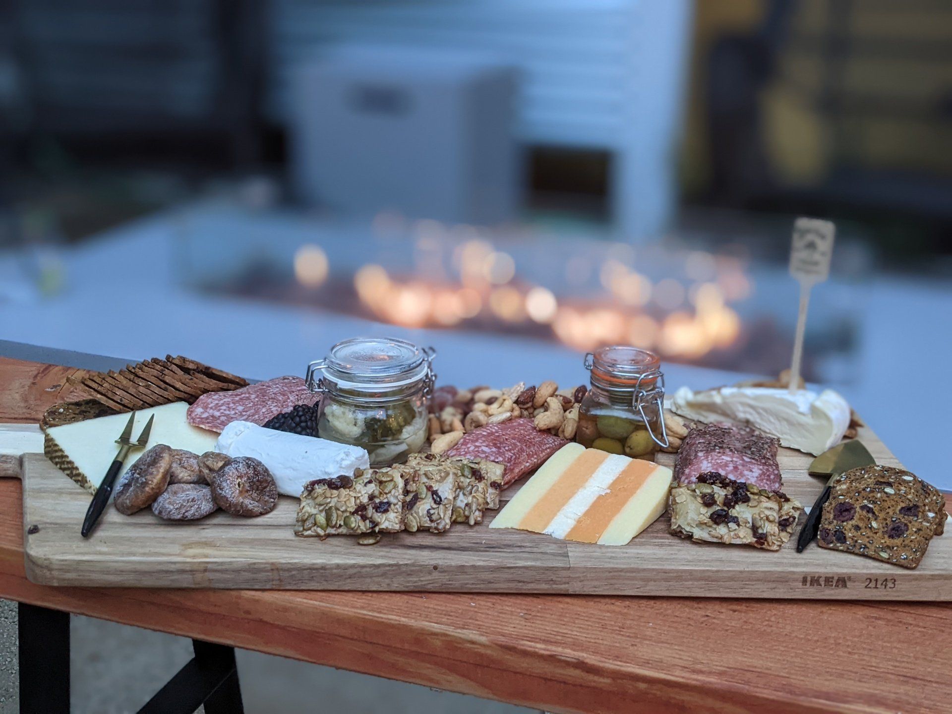 There is a fire pit in the background and a wooden cutting board with a variety of food on it.