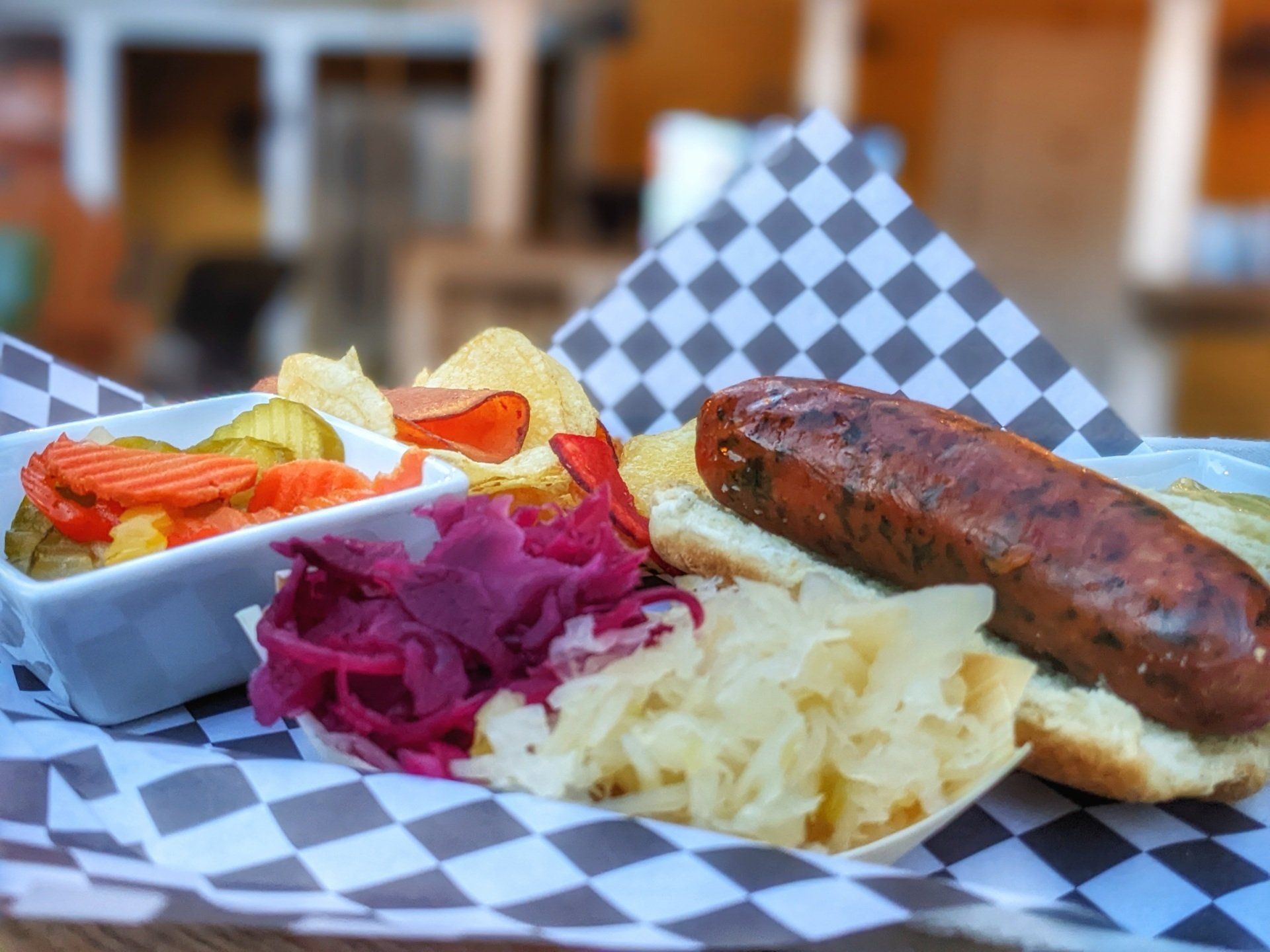 A sausage sandwich with sauerkraut and potato chips on a checkered paper on a table.