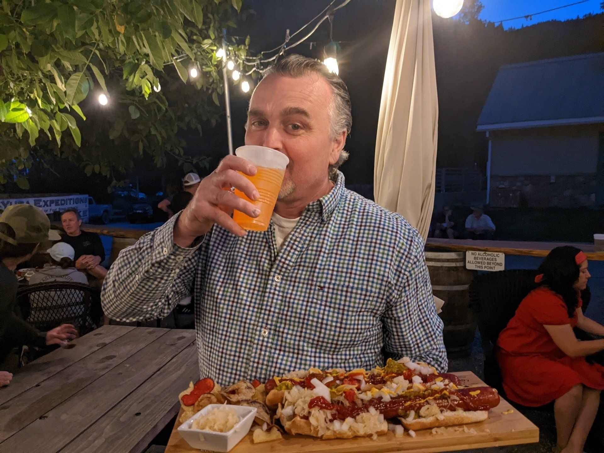 A man is drinking a beer and eating a hot dog on a cutting board.