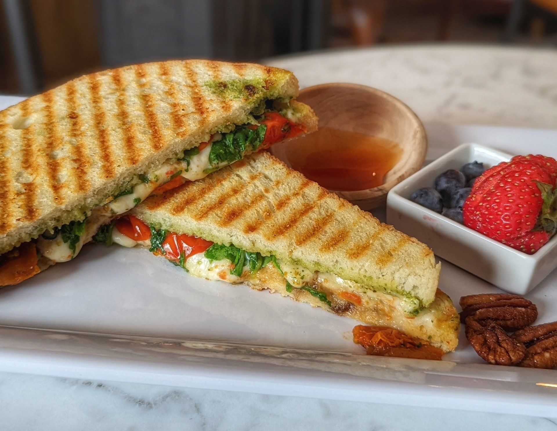 A sandwich is cut in half on a white plate next to a bowl of strawberries and blueberries.
