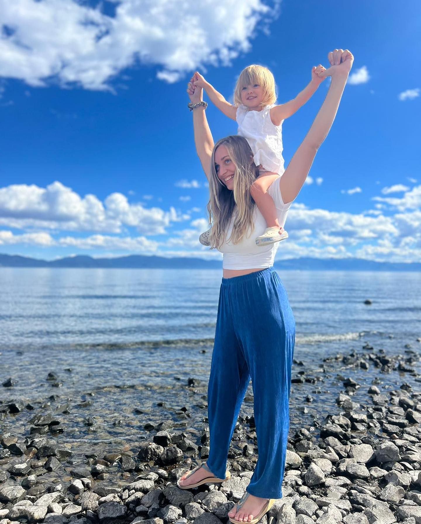 A woman is carrying a little girl on her shoulders on the beach.
