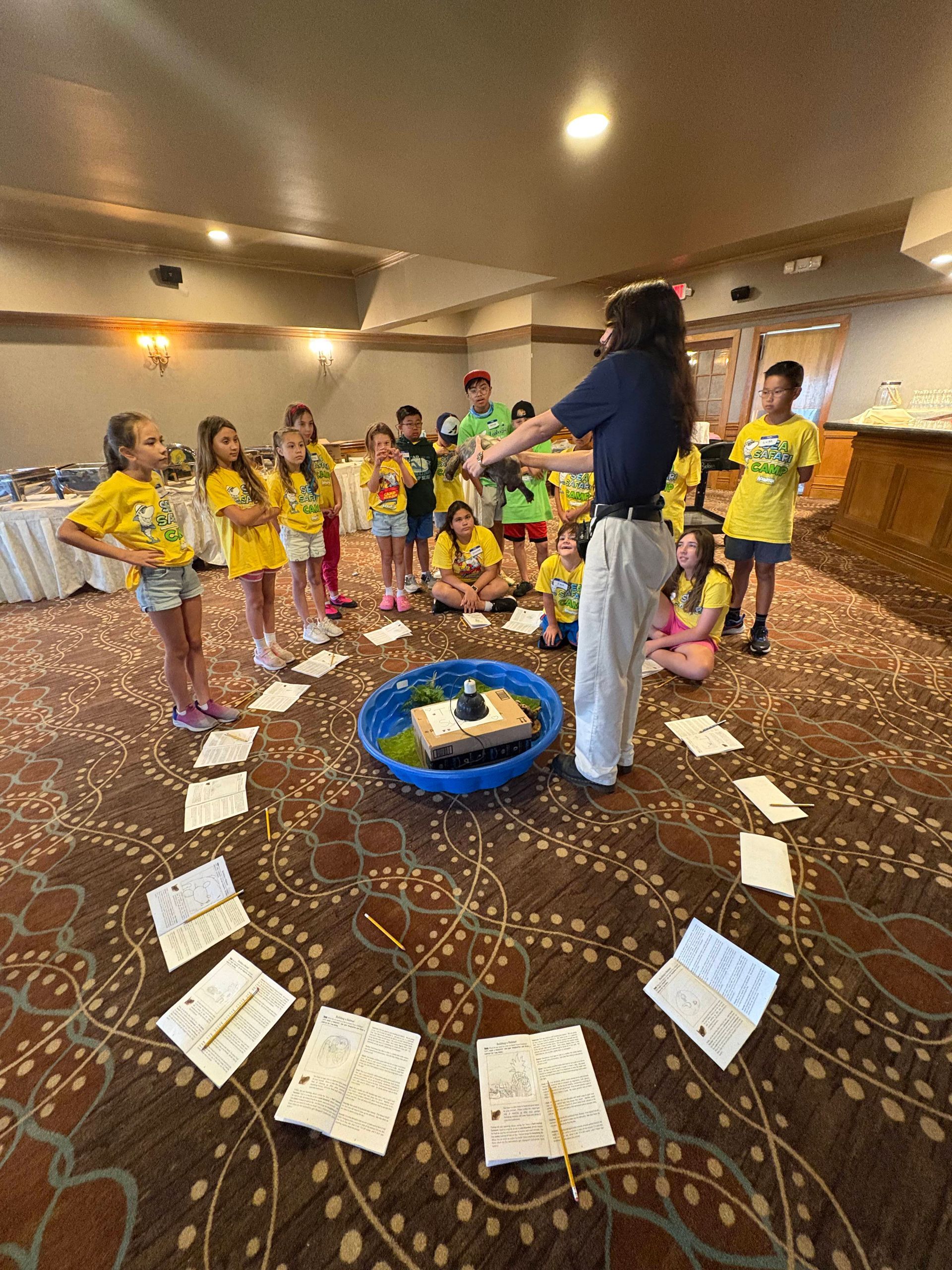 Children in yellow shirts gather around a woman and a small pool on a carpeted floor.