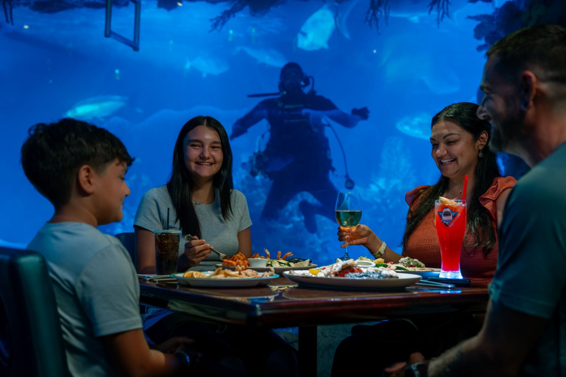 A family eats at an underwater restaurant while a scuba diver swims in the background aquarium tank.