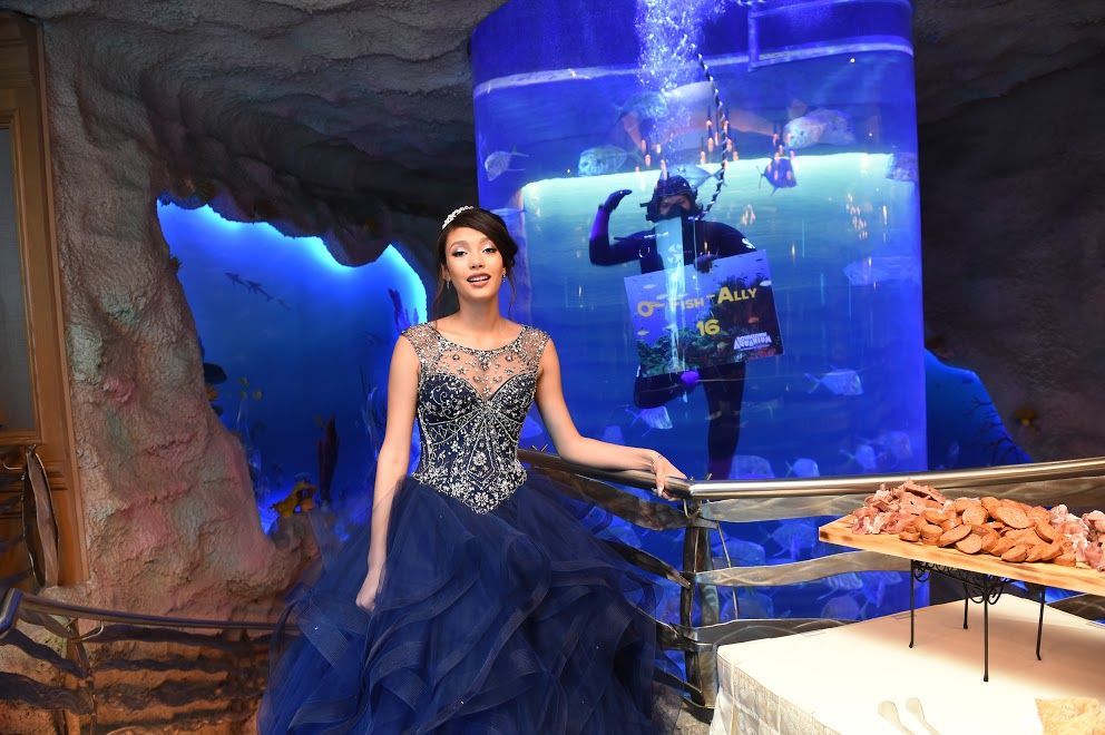 Woman in blue gown poses near a large aquarium with a scuba diver inside.