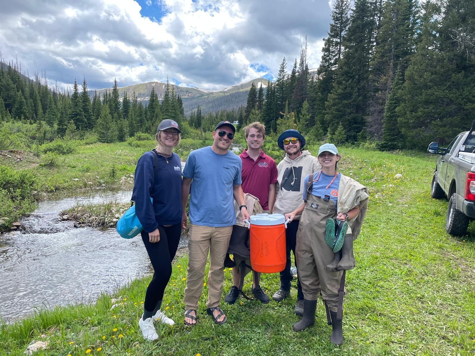 A group of people standing next to a river holding an orange cooler.