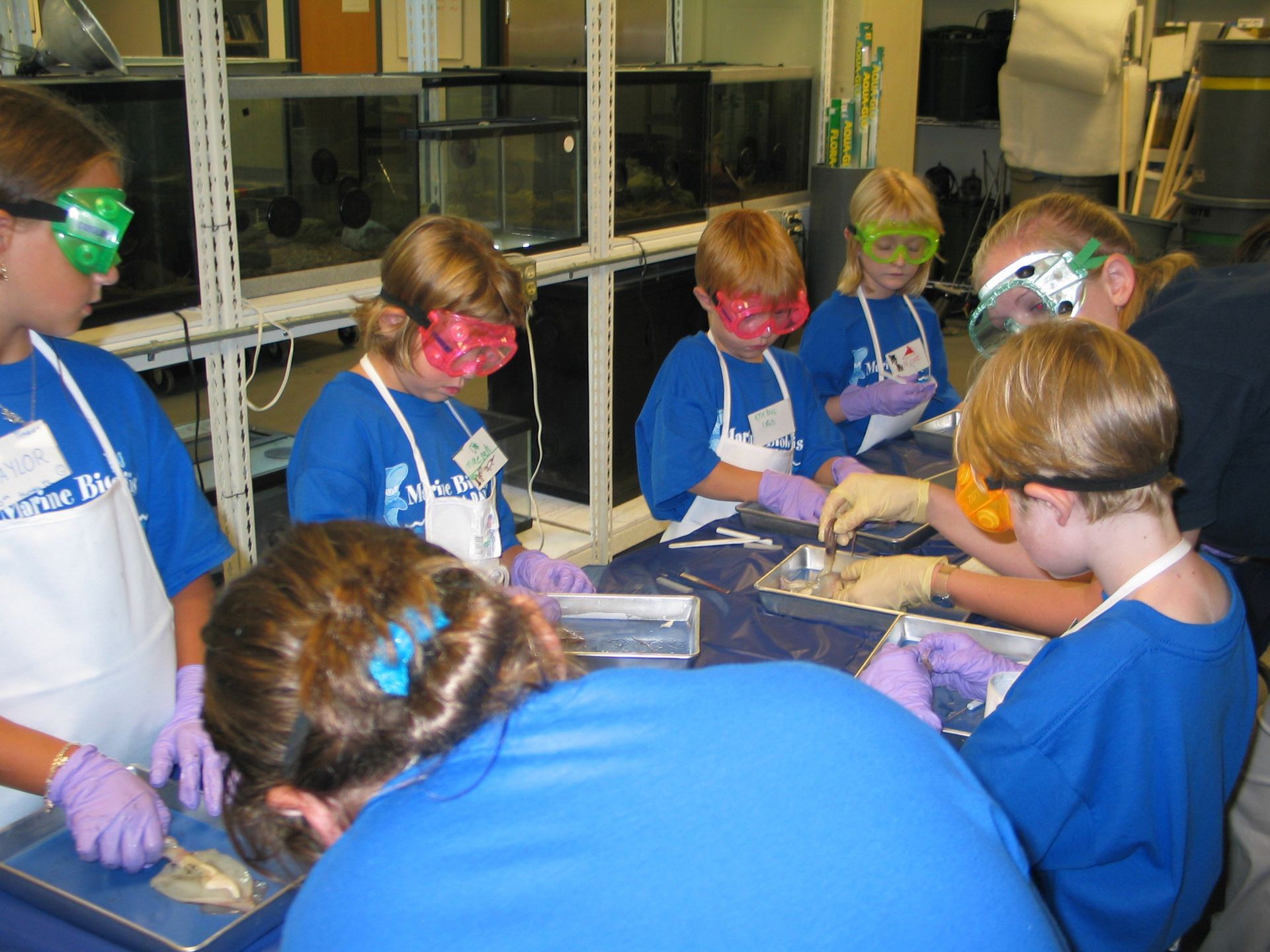 A group of children wearing safety goggles and aprons are working in a lab