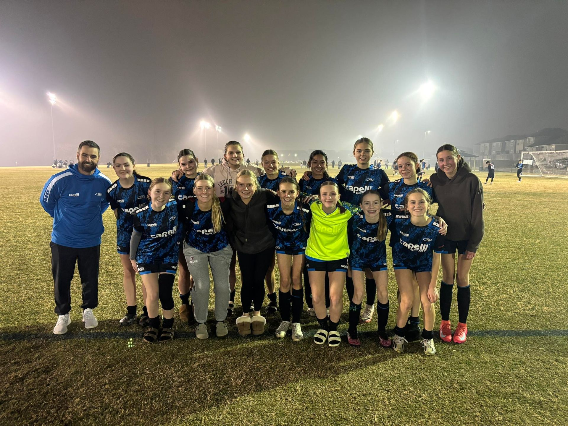 Soccer team poses on field at night under stadium lights. Players wear blue and black jerseys, some smile.