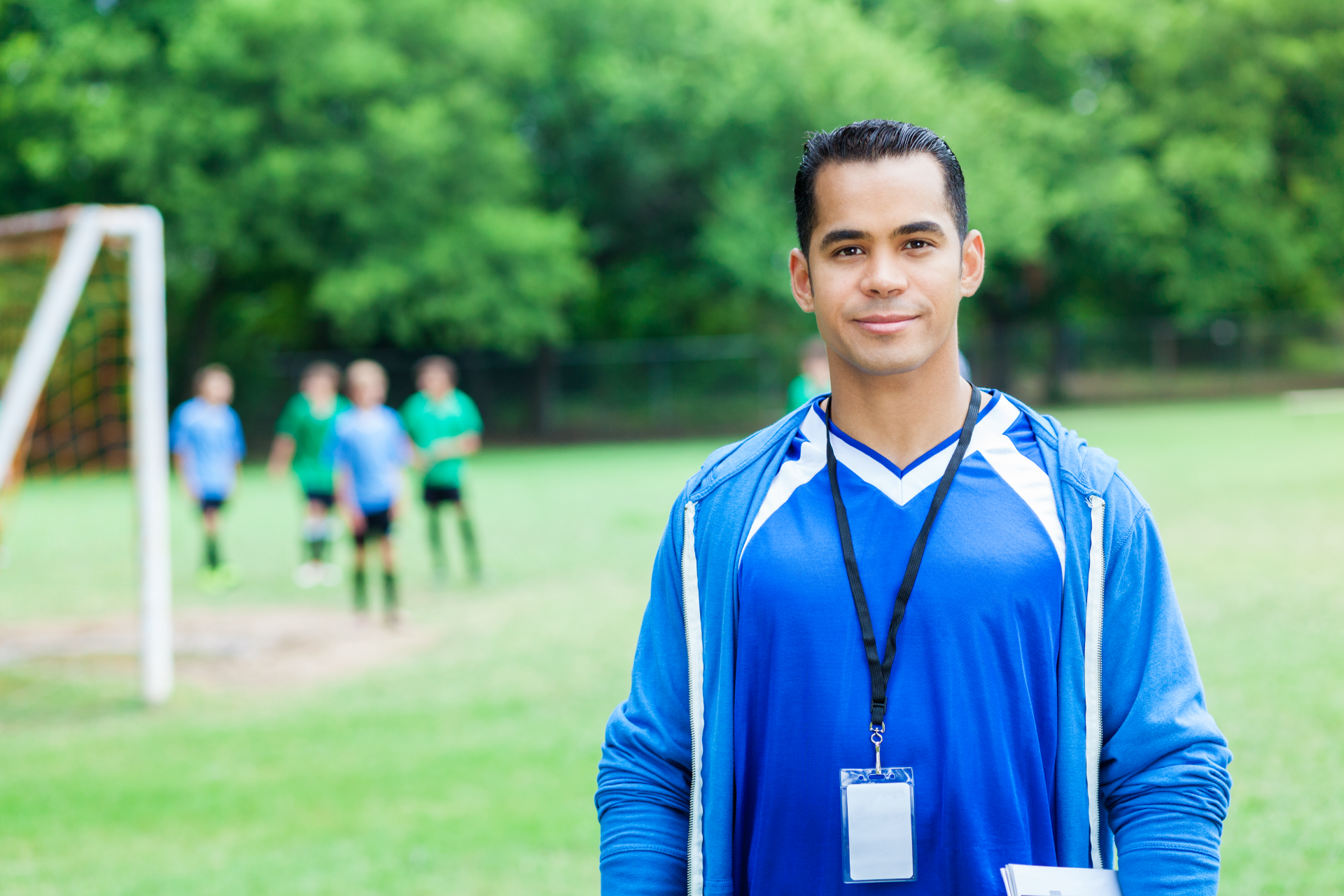 Soccer coach wearing blue stands on field, looking towards the camera; players in background.