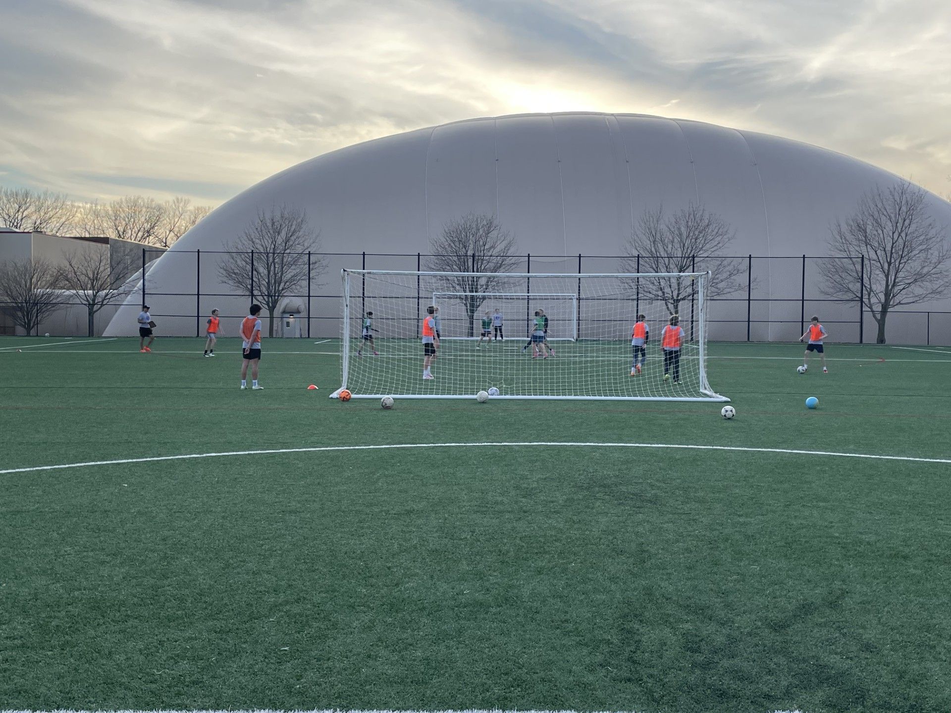 Soccer players in orange practice shooting goals on a field, dome in the background.