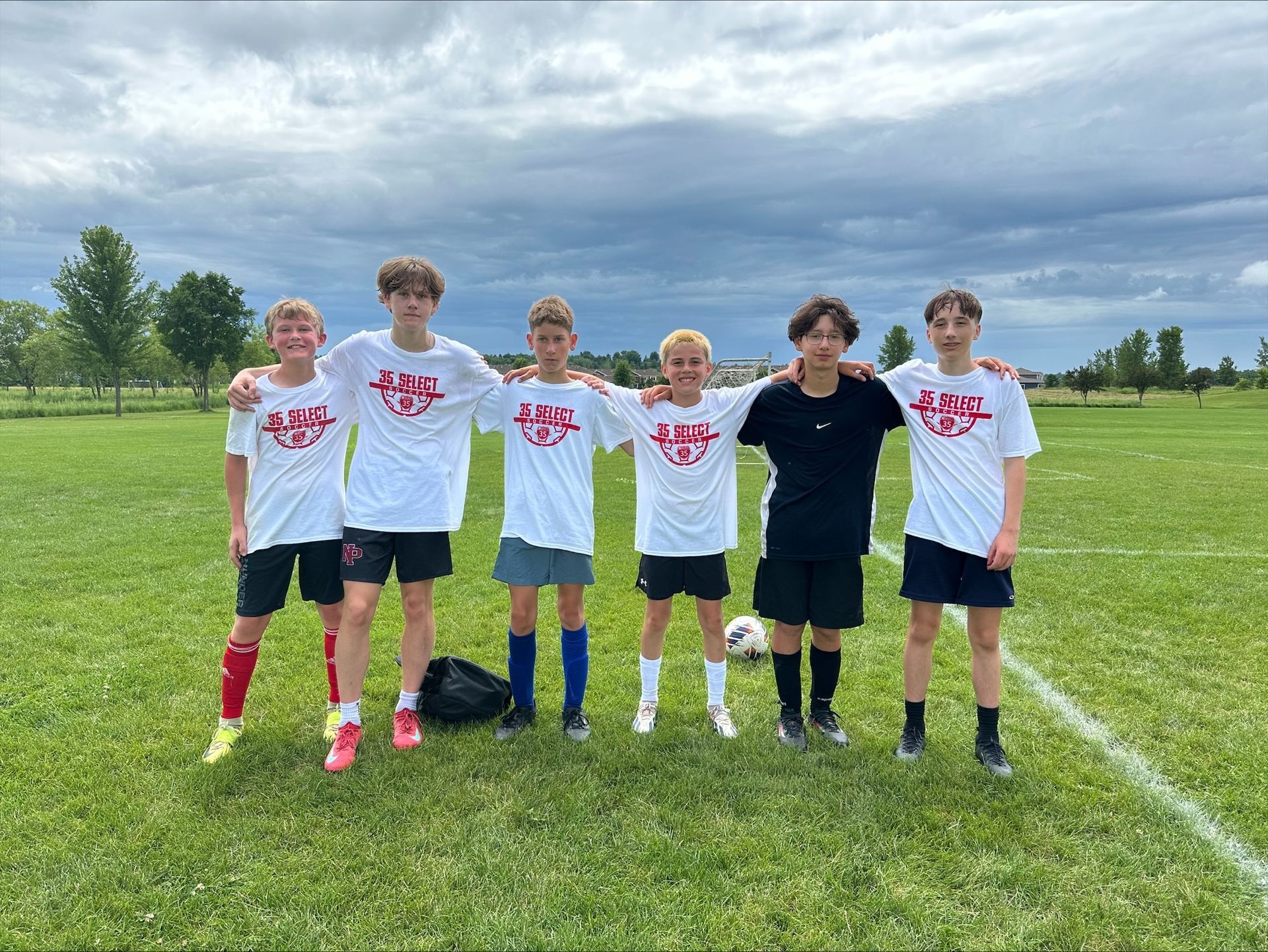 Soccer team of six youth stand on a grass field, arms around each other, wearing matching white shirts and black shorts.