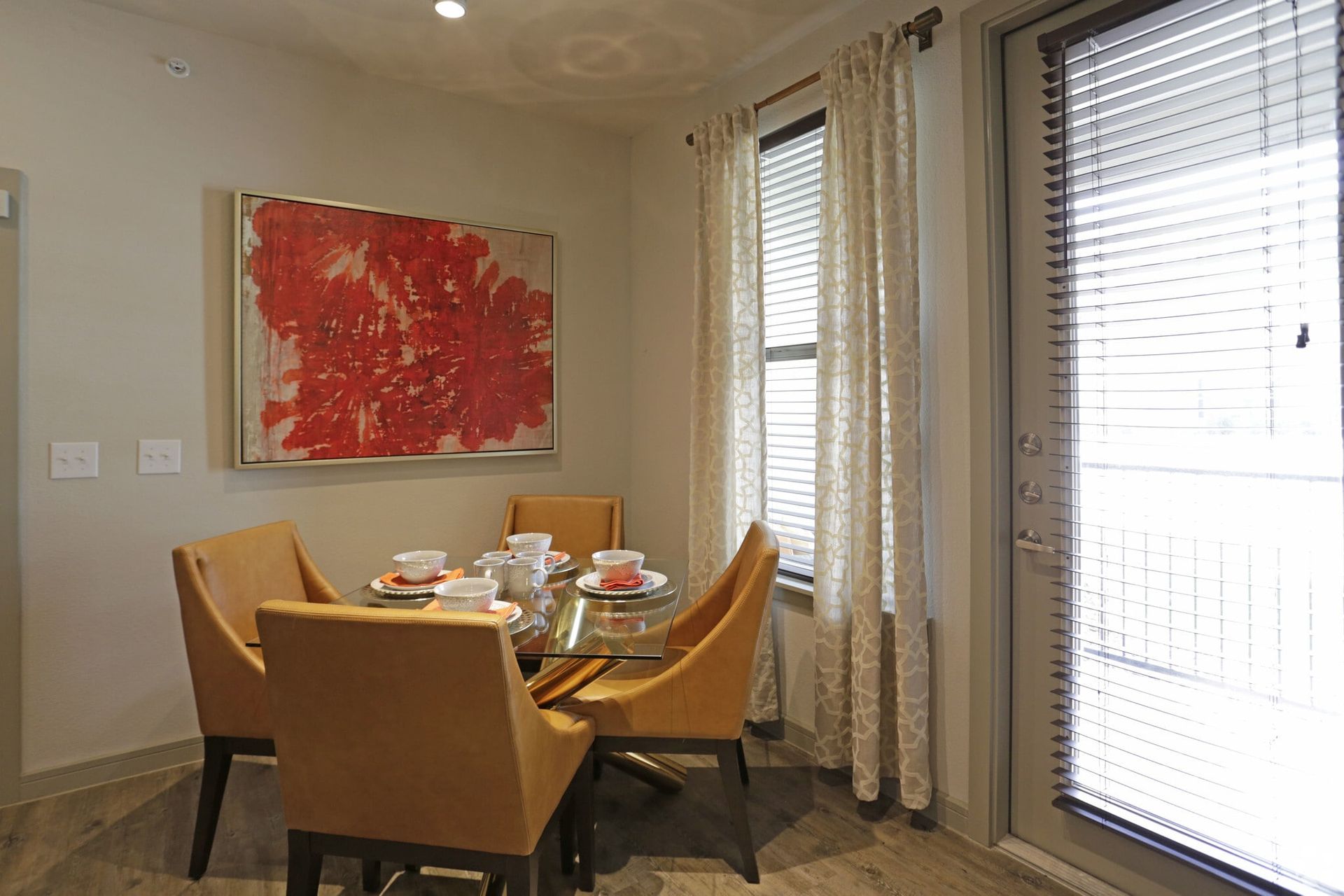 Dining area with glass table, four tan chairs, red abstract art, and window blinds.