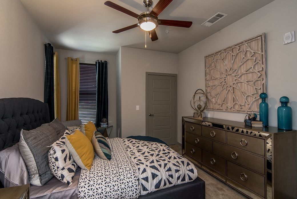 Bedroom in an apartment with a gray upholstered bed, dresser, ceiling fan, and decorative wall art.