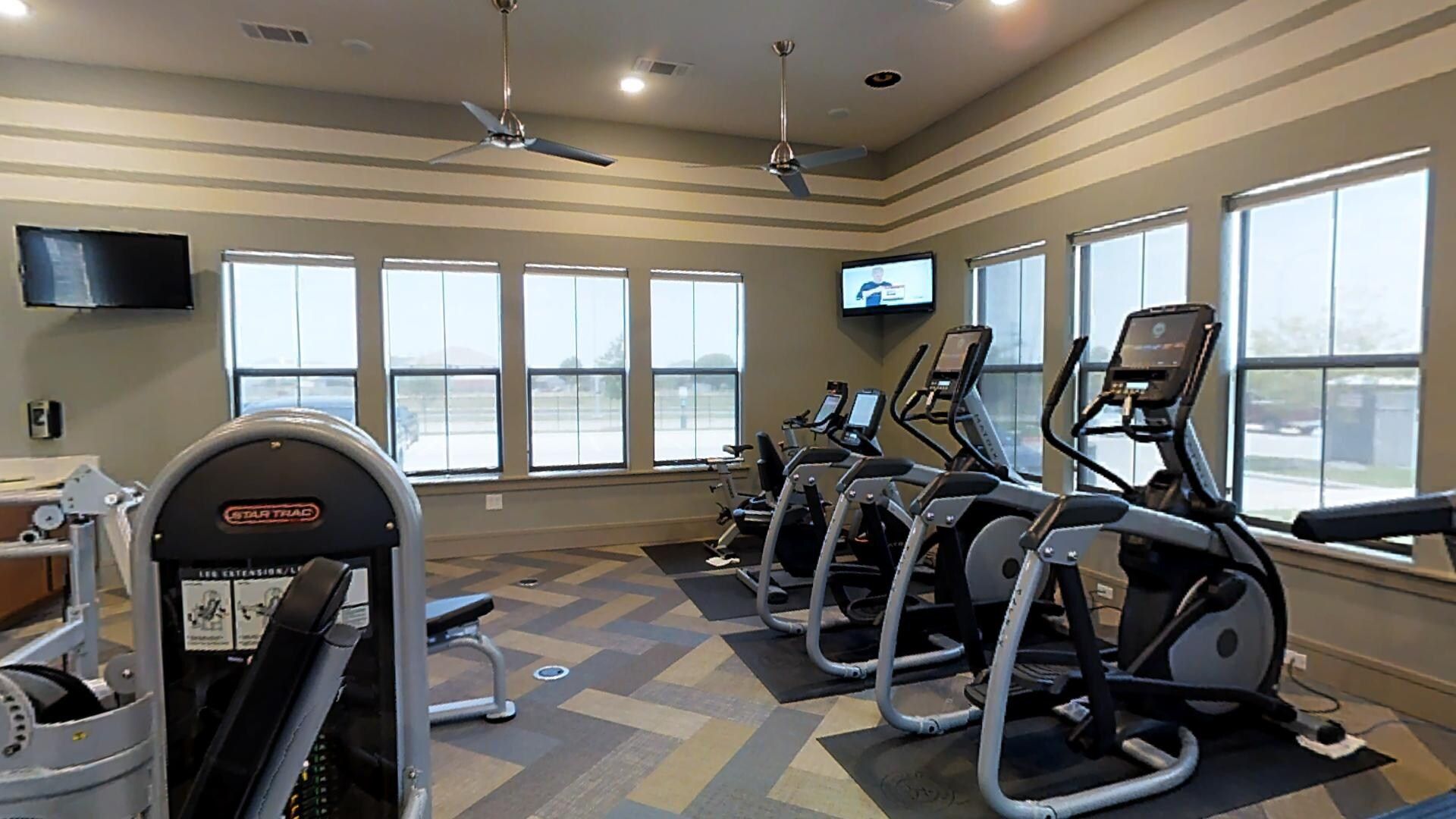 Indoor gym with treadmills and ellipticals by large windows.