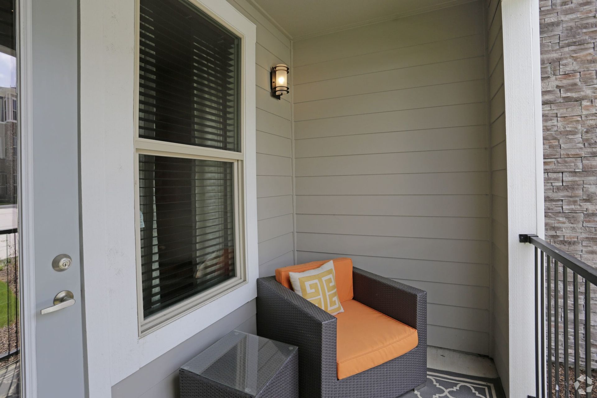 Apartment balcony with a grey wicker chair, orange cushions, and a small glass table.