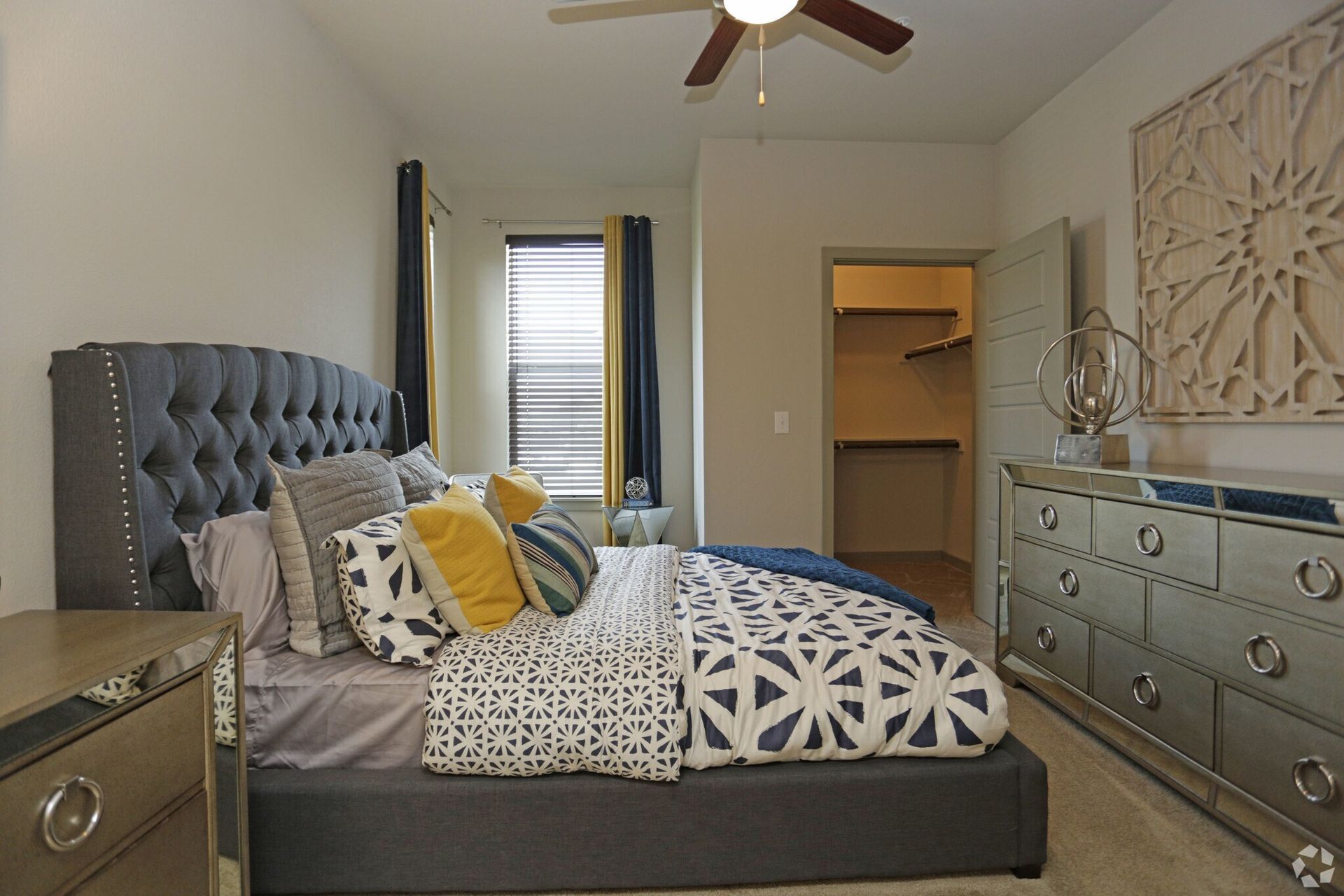 Bedroom in a gray tufted bed with a mirrored dresser and closet, window with blue and yellow curtains.