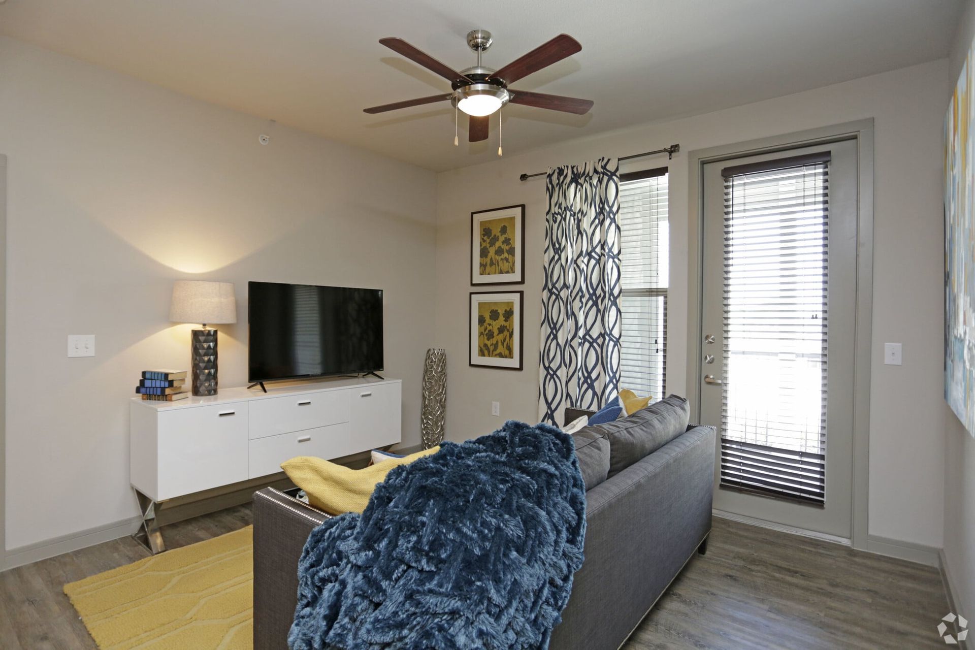 Living room with gray sofa, white media console, flat-screen TV, ceiling fan, and patterned curtains.