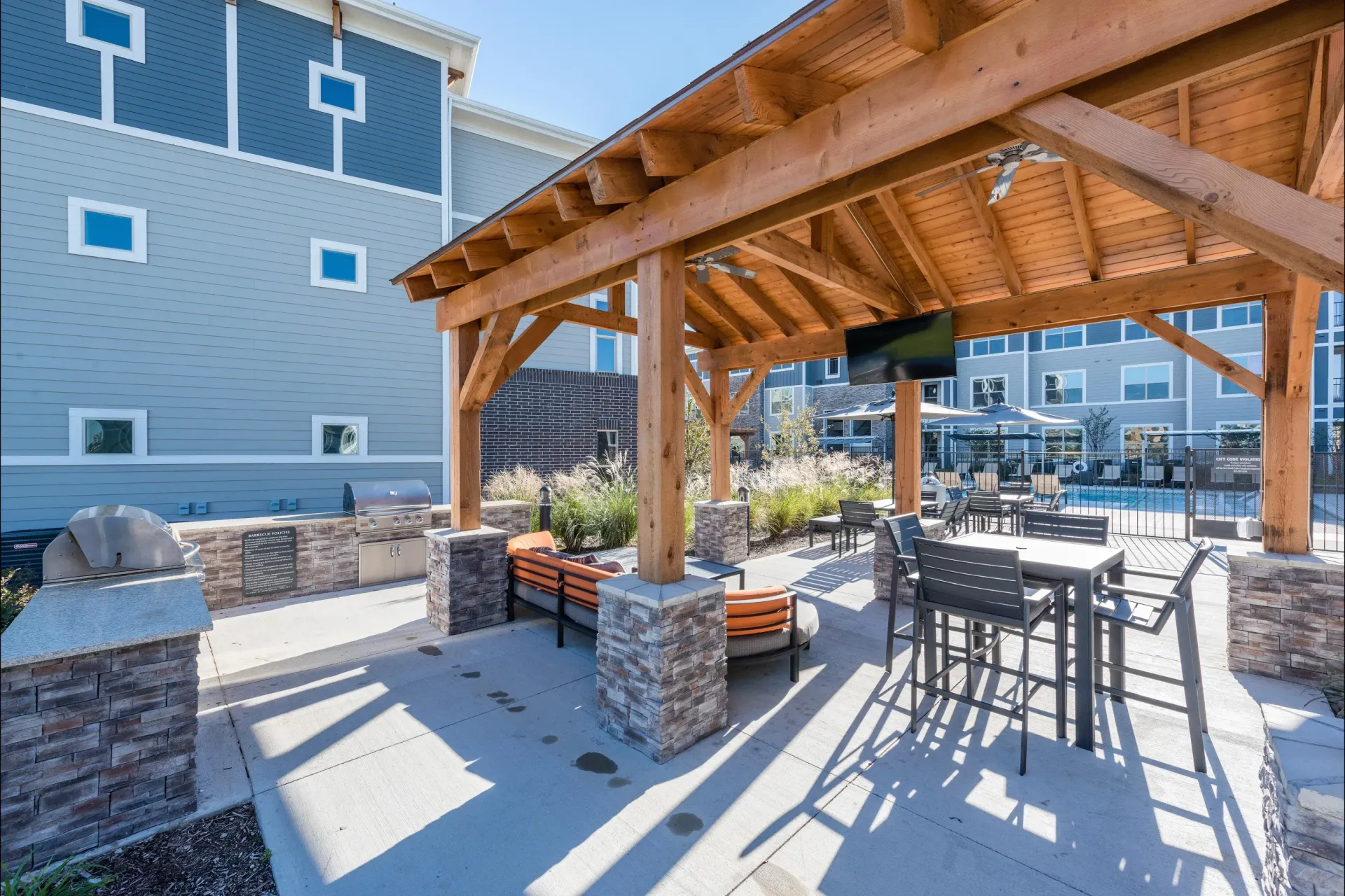 Outdoor community amenity with a wooden pavilion, seating, grills, and a pool in the background.