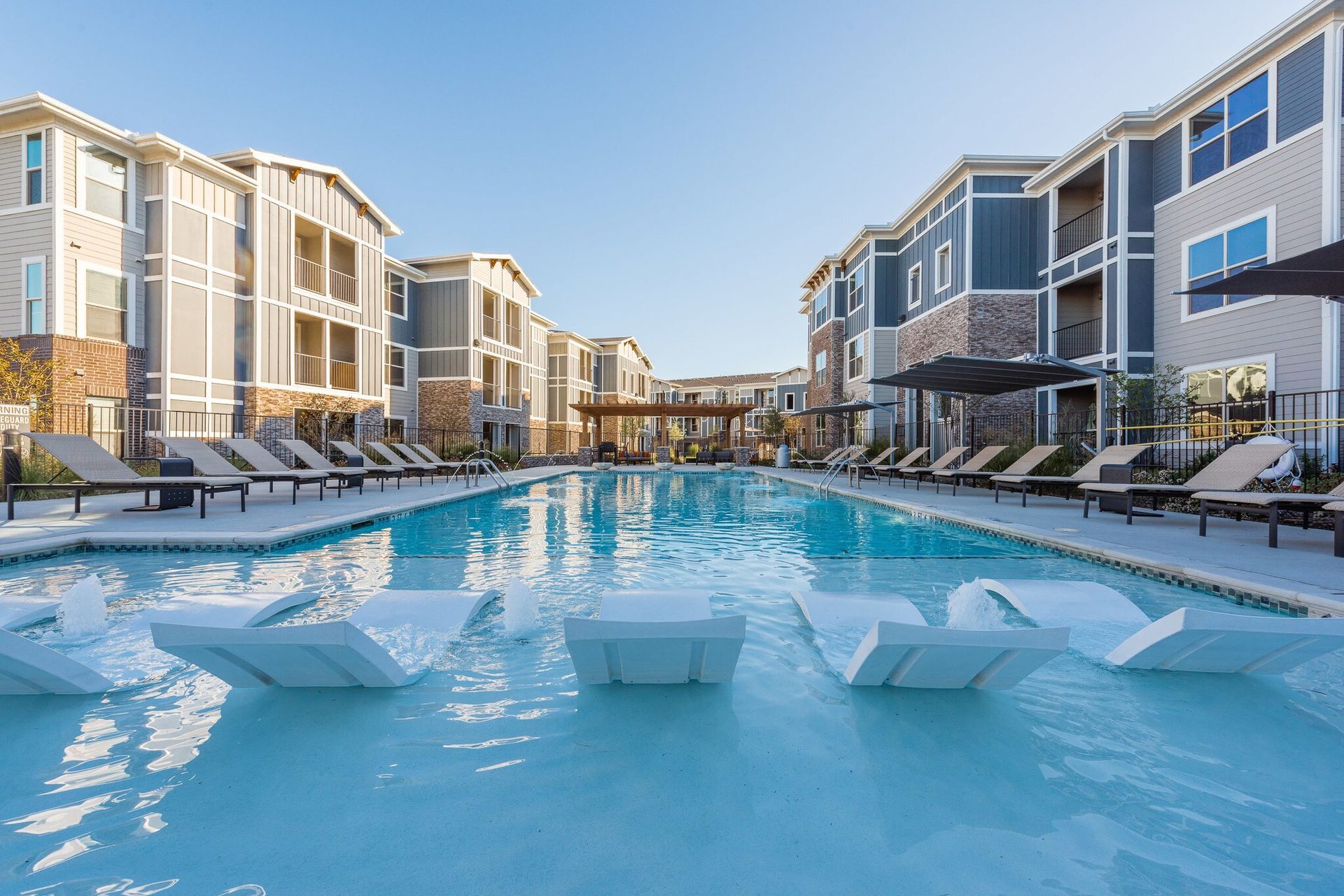 Outdoor pool at a modern apartment complex with lounge chairs along the deck and surrounding buildings.
