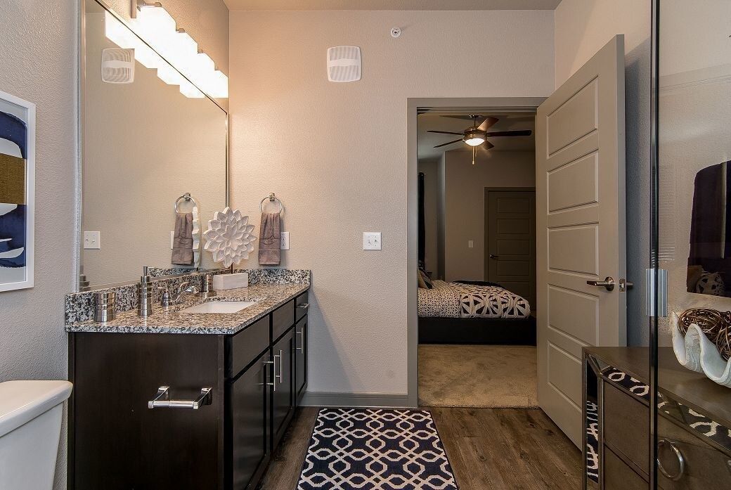 Bathroom with a double-sink granite vanity, large mirror, towels, and view into a bedroom.