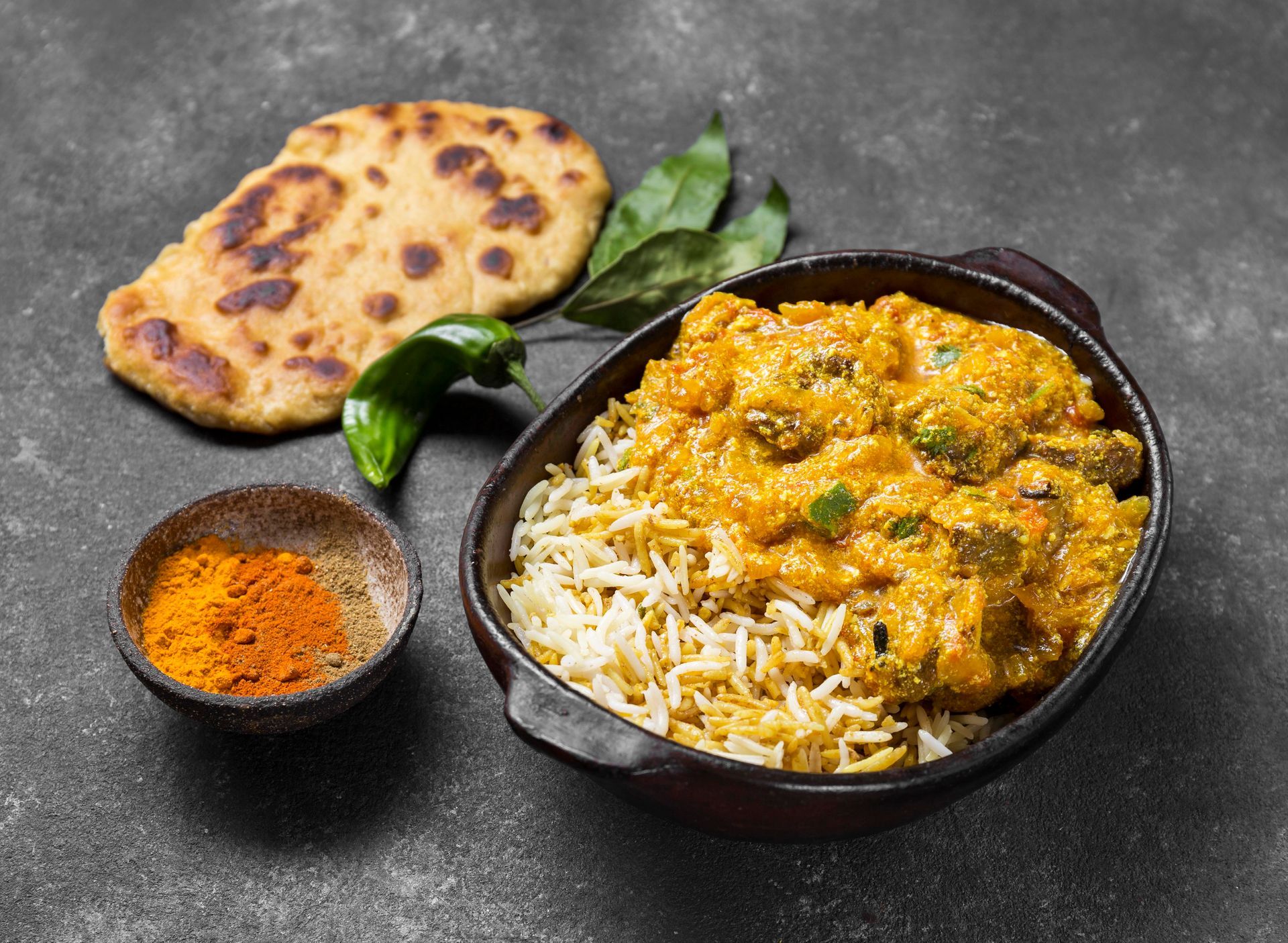 A bowl of curry over rice next to a piece of flatbread, some green leaves, and a small bowl of yellow spice powder.
