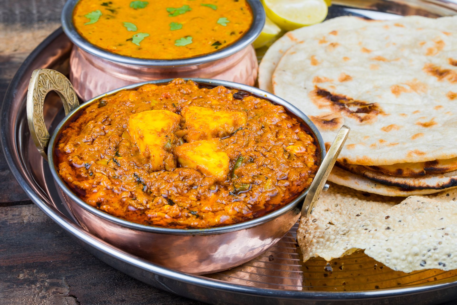 A metal platter holding two copper bowls of Indian curry, flatbread, and a papadum with lemon wedges.