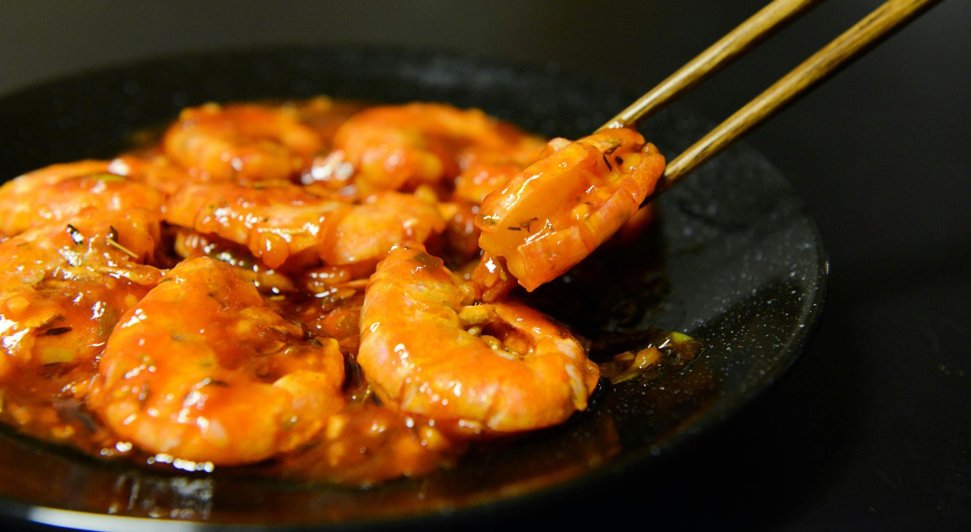 A pair of chopsticks lifts a cooked shrimp from a plate of saucy, glazed shrimp against a dark background.