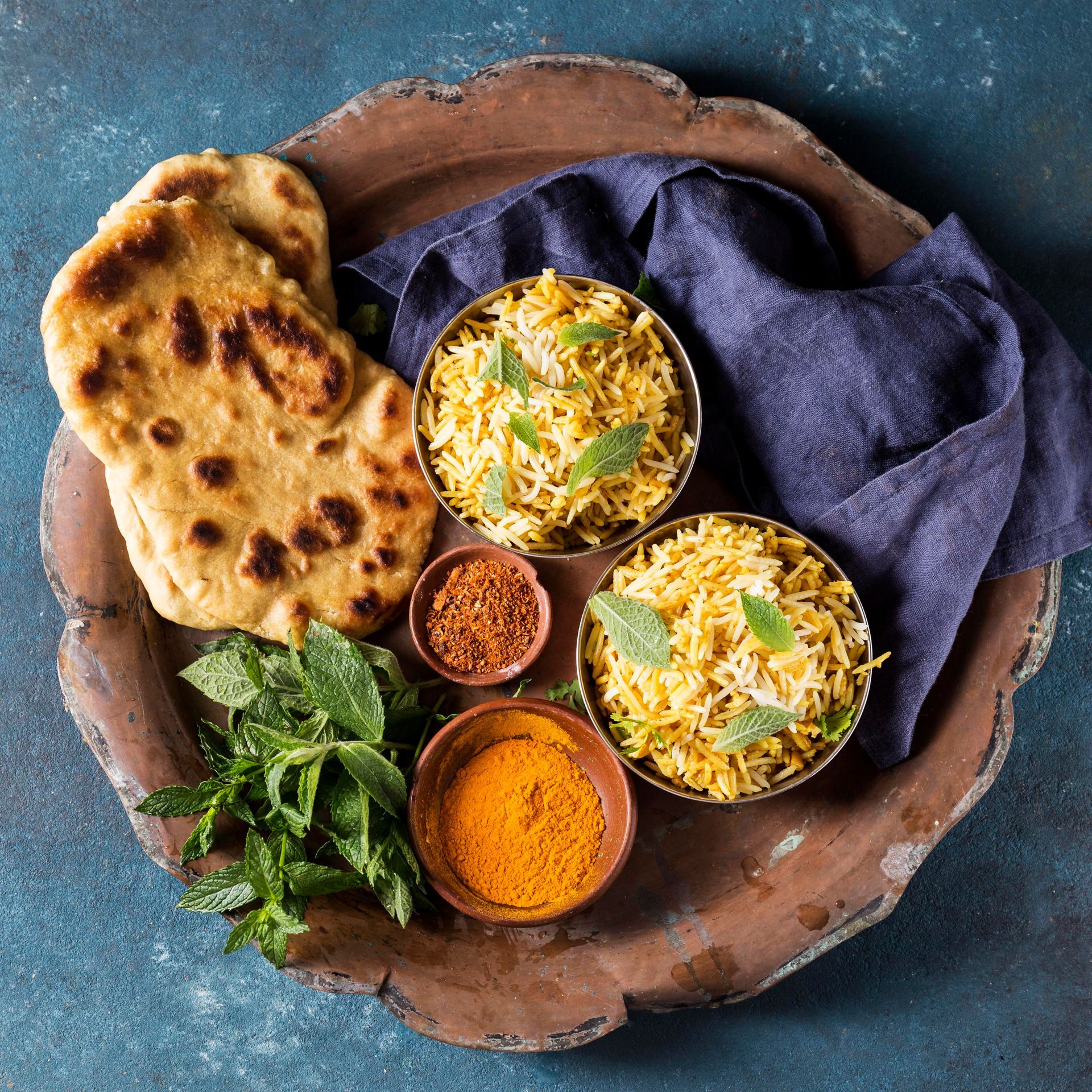 Two bowls of yellow rice, flatbread, fresh mint, and small bowls of spices on a rustic tray against a blue background.
