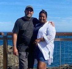 A man and a woman are posing for a picture in front of the ocean.