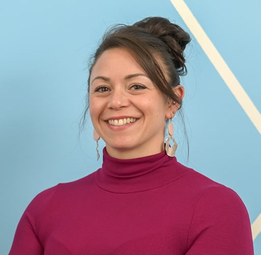 A woman wearing a purple turtleneck and earrings is smiling for the camera.