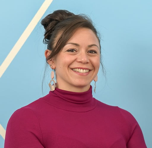 A woman wearing a purple turtleneck and earrings smiles for the camera