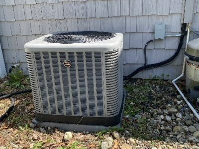 Air conditioning unit outside a house with gray siding. Black electrical conduit and gray control box.
